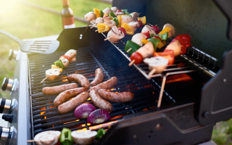 Das Bild zeigt einen geöffneten Gasgrill mit Würstchen, Gemüsespießen und Zwiebeln auf dem Rost. Die sichtbaren Flammen und das Grillgut vermitteln eine lebendige, sommerliche Grillsituation im Freien.