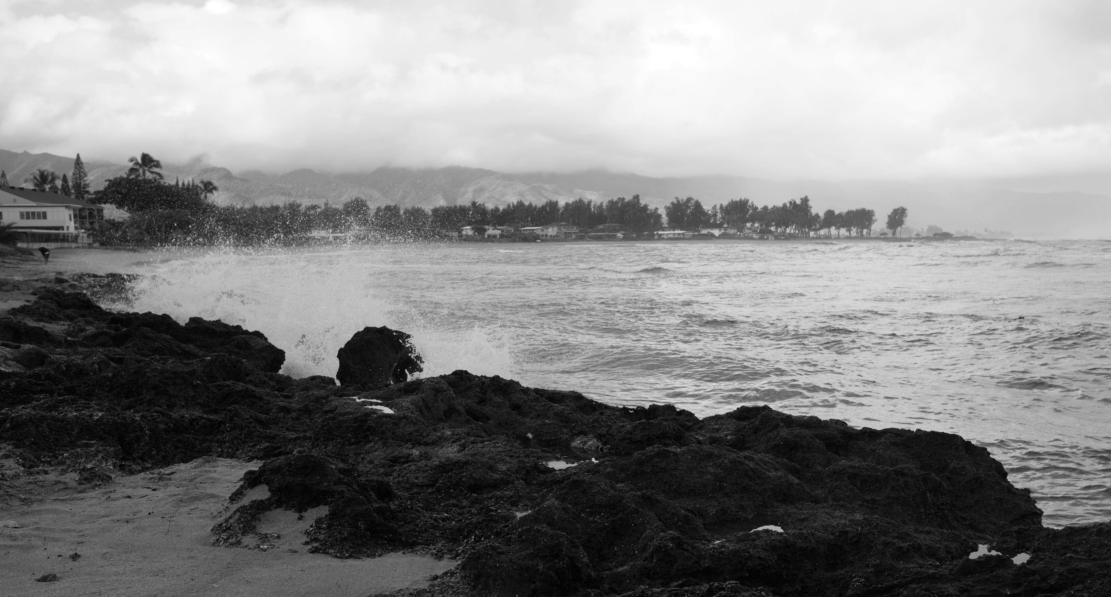 Waves softly crashing on the black rocks. A beach on the left leading to a peninsula of houses on the other side — a man is walking away from the camera a medium distance away. A cloudy mountain ridgeline in the far distance.