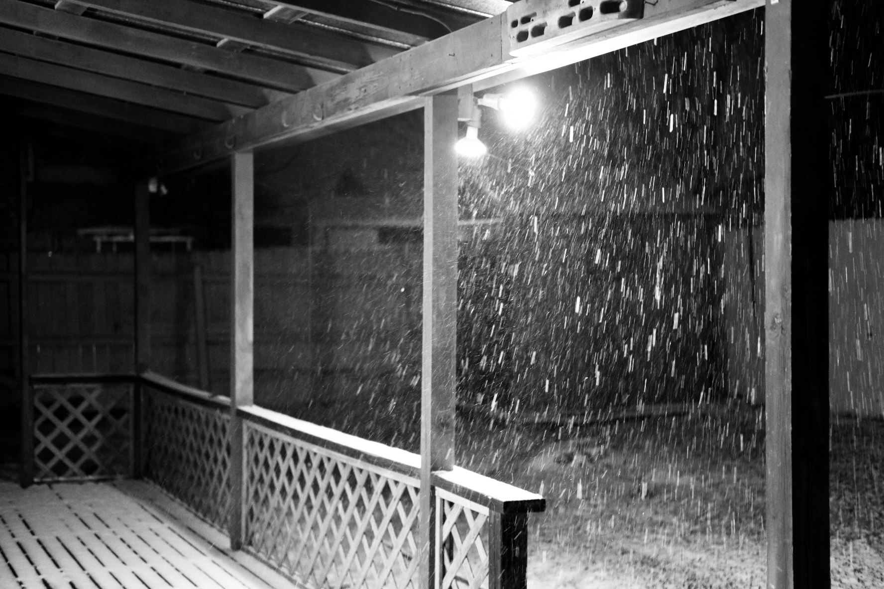 Light snow flurries next to our backyard flood lights. The deck covered in a thin layer of snow under the awning. A small shed sit in the corner of the yard.