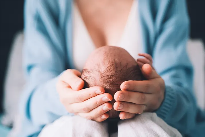 Femeie așezată pe o canapea, care ține  în poală un bebeluș, în timp ce îi susține căpșorul cu mâinile.
