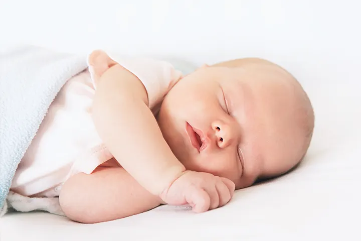 Bebeluș care stă întins pe o parte, cu mânuțele lângă urechea  care atinge patul.
