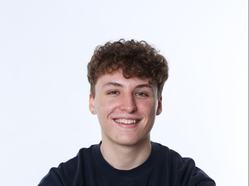 Idris schaut in die Kamera. In einem professionellem Fotostudio sitzt er und trägt ein schwarzes T-Shirt. Er hat kurze braune lockige Haare und lächelt freundlich.