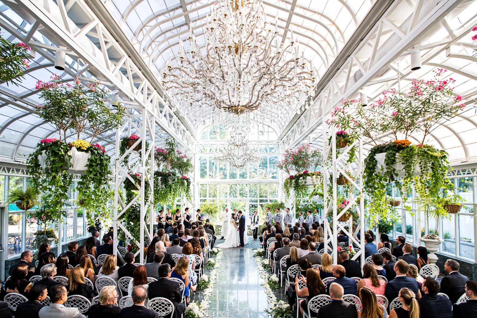 Wedding ceremony in an elegant glass conservatory with crystal chandeliers, lush plants, and guests seated in rows.