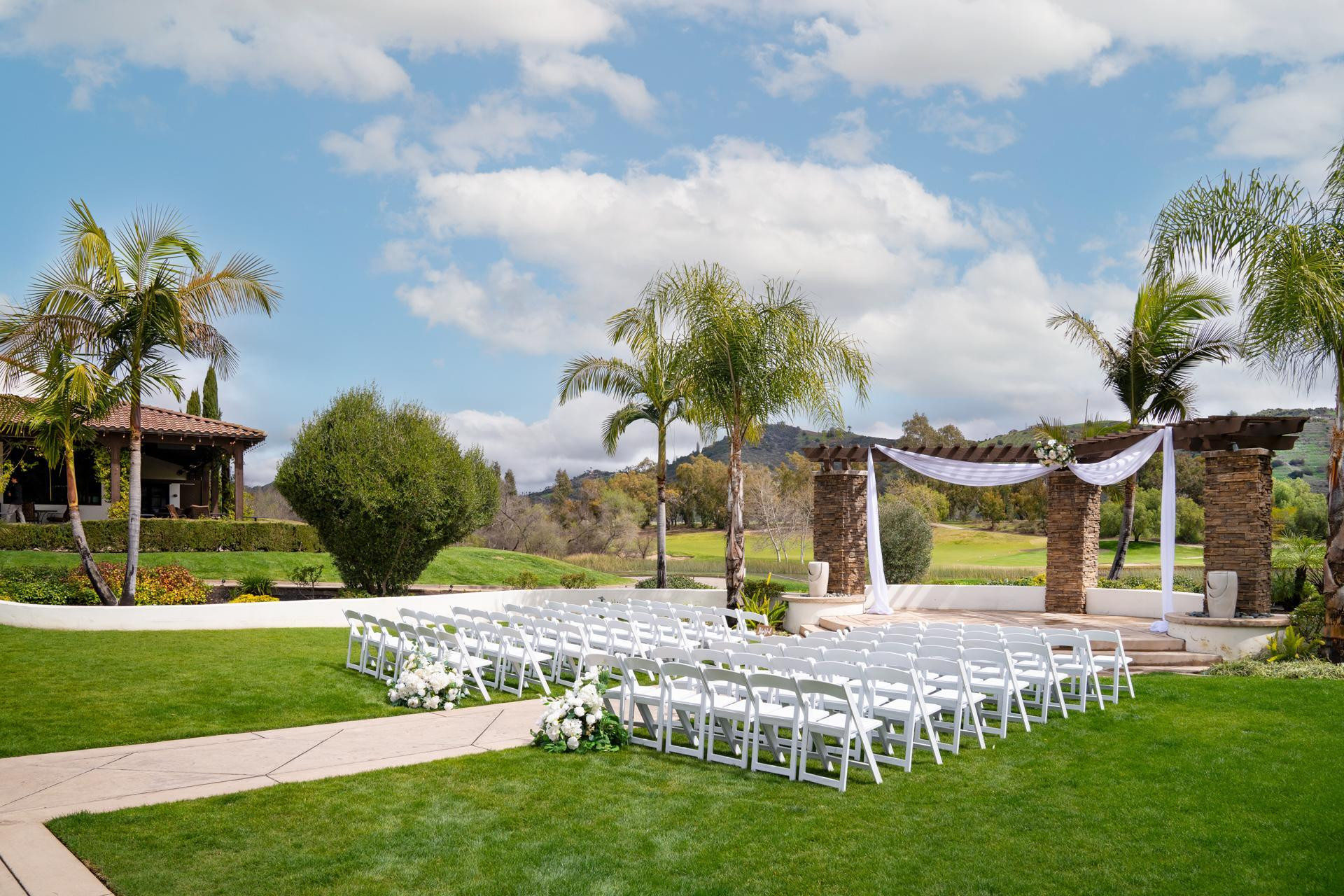 Outdoor wedding venue with stone pergola draped in white fabric, rows of white chairs, and scenic golf course backdrop.