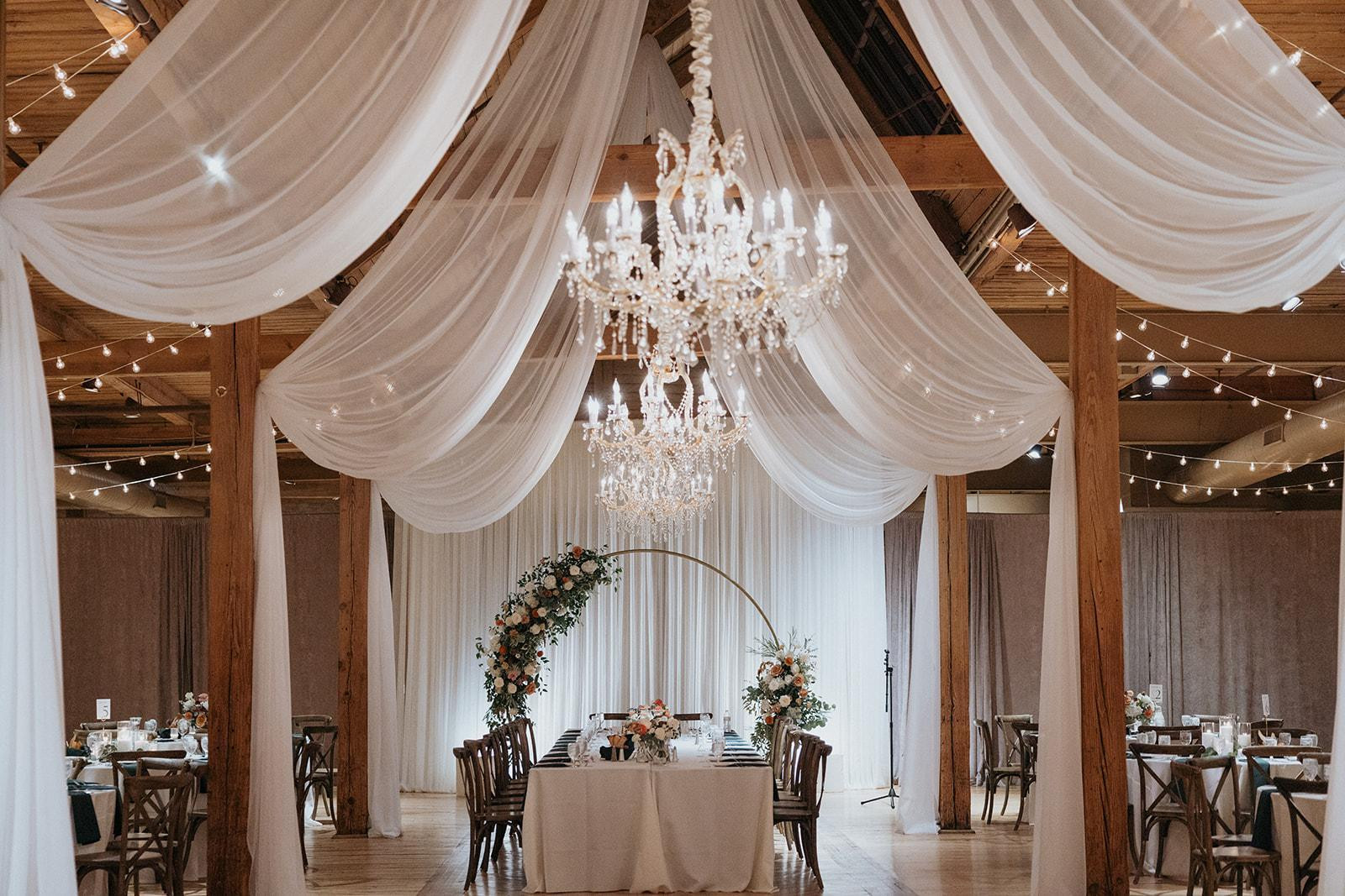 Couple kissing in brick venue with glass ceiling, chandeliers, and flowing veil against backdrop of wooden chairs and white flowers.