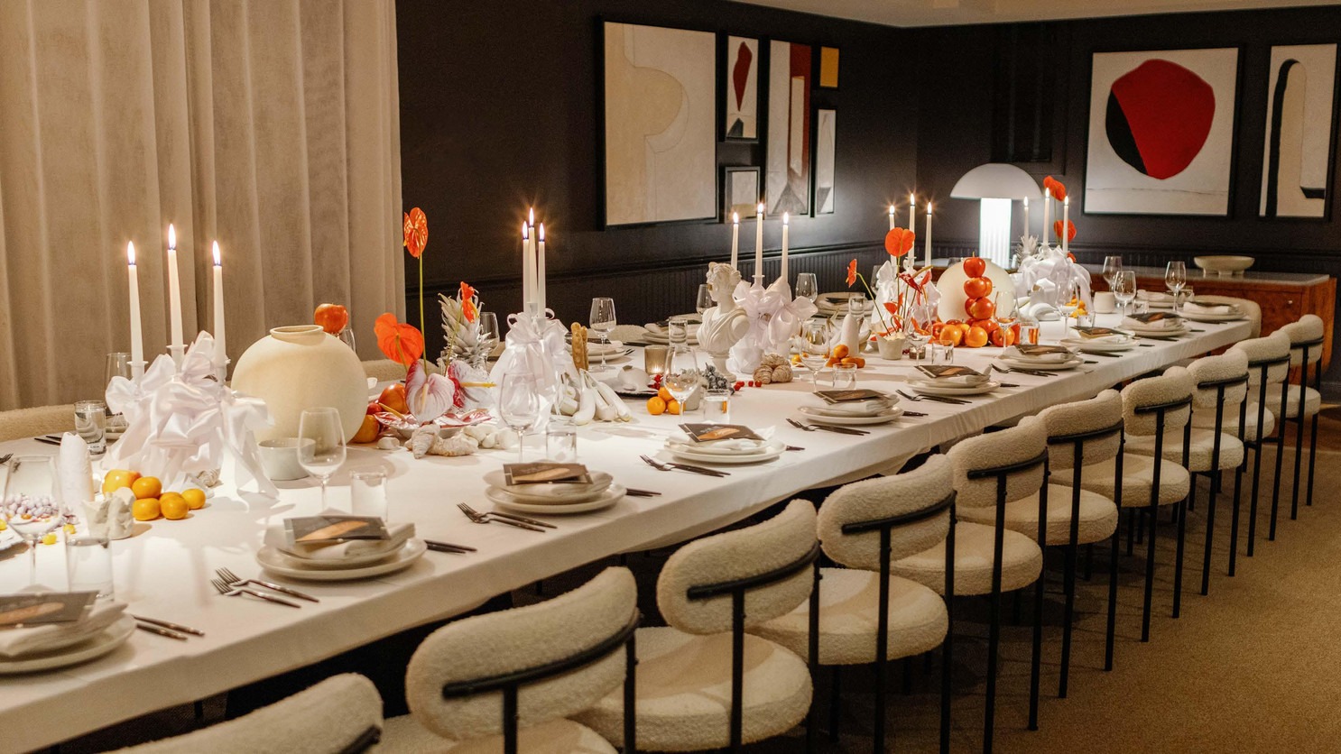Elegant dining table with white tablecloth, cream chairs, candles, and orange accents against dark walls with modern artwork.