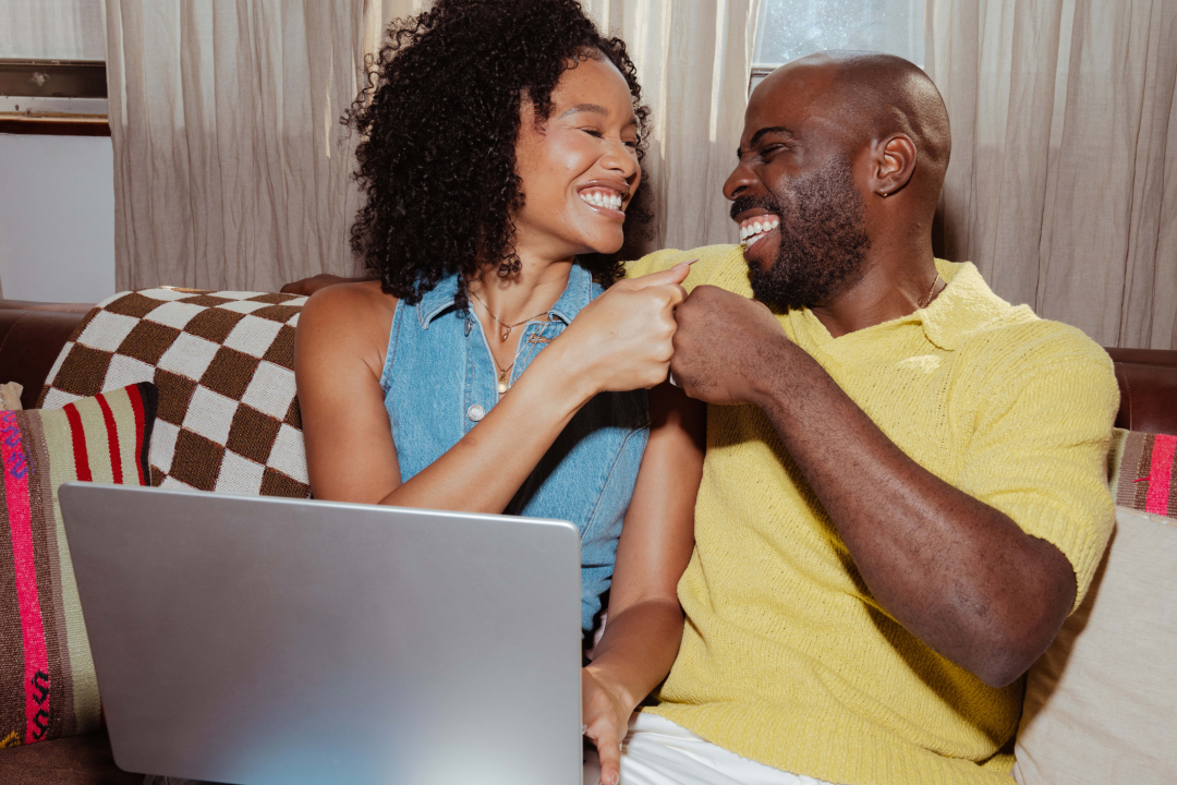 Smiling couple sharing a joyful moment on couch with laptop, wearing blue and yellow tops against patterned pillows.