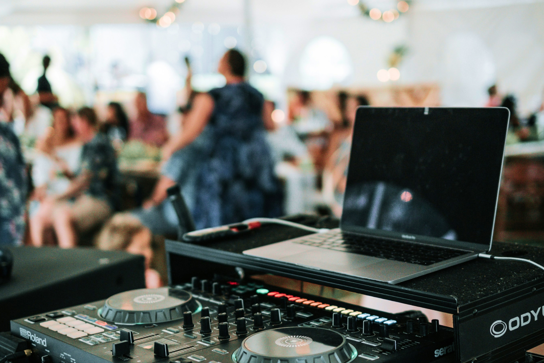 DJ equipment with laptop and mixer at an event with blurred guests in the background and soft lighting.