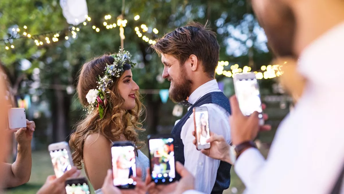 Guests with smartphones taking photo of bride and groom at wedding reception outside in the backyard