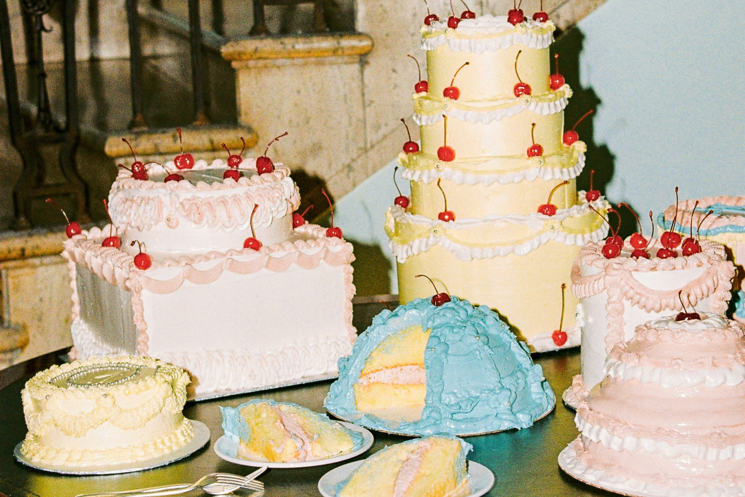 Vintage-style display of decorated cakes with cherries, including a tall yellow tiered cake and blue dome cake with slices.