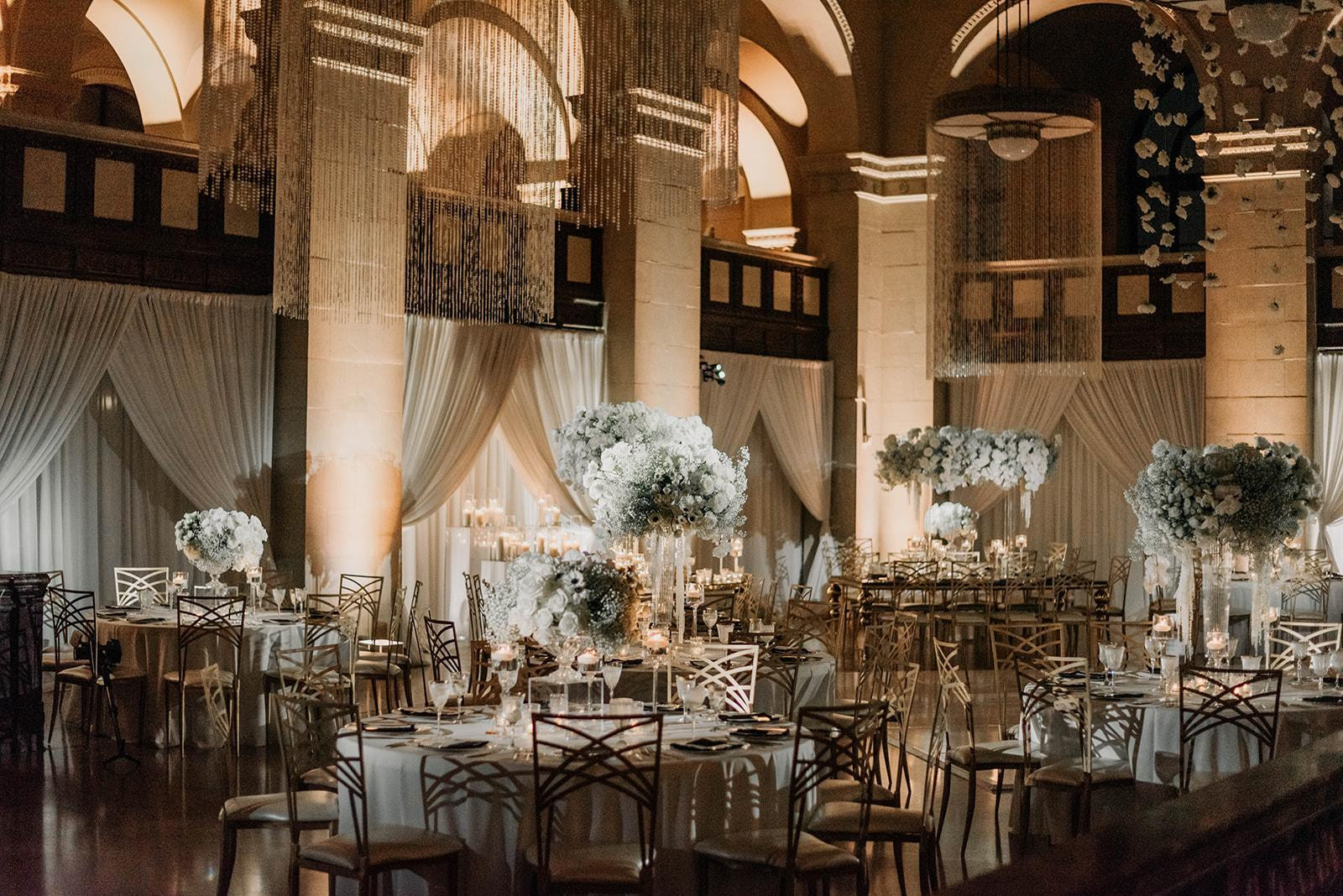 Elegant ballroom wedding reception with crystal chandeliers, white floral centerpieces, and gold chairs in warm ambient lighting.