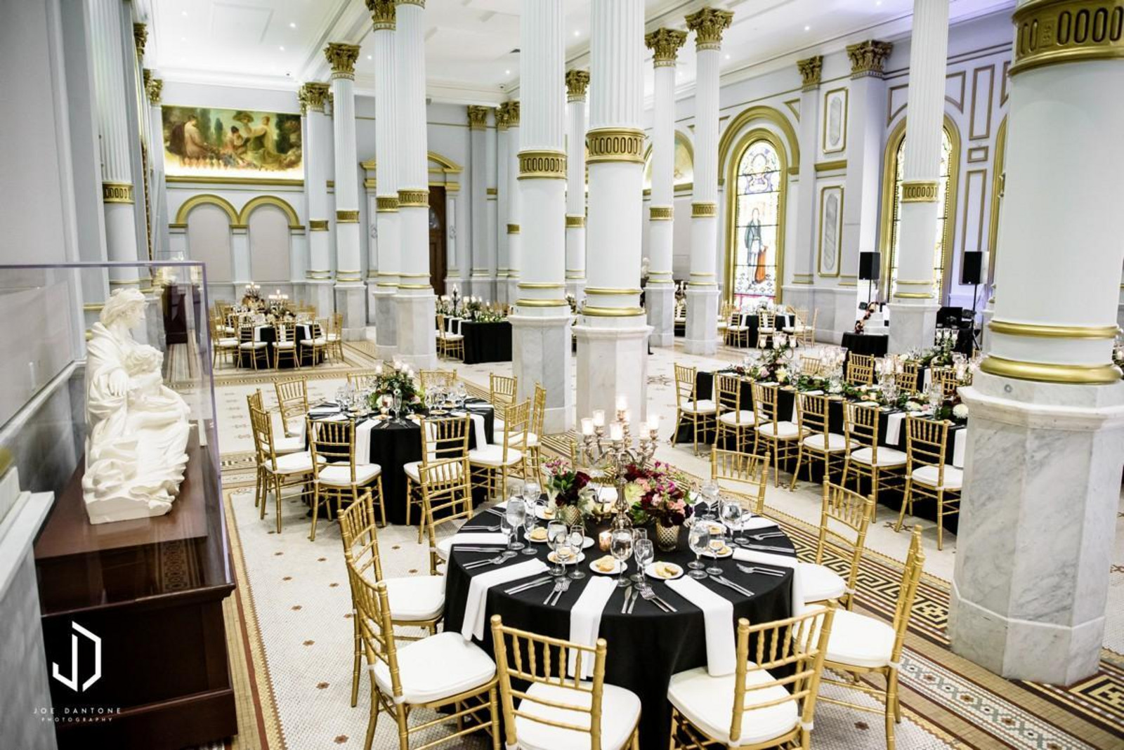 Elegant banquet hall with white columns, gold accents, and round tables with black tablecloths and gold chairs set for formal dining.