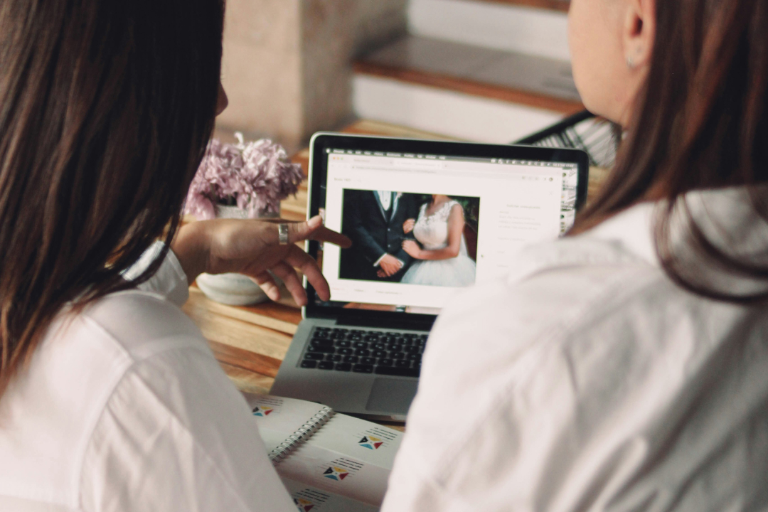 Two people in white shirts looking at wedding photo on laptop screen, with lilac flowers nearby on wooden table.