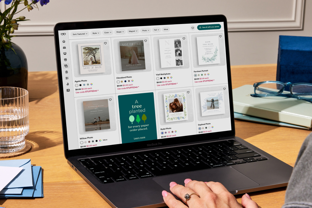 Laptop displaying wedding Zola save-the-date card designs on a wooden desk with flowers and glasses nearby.