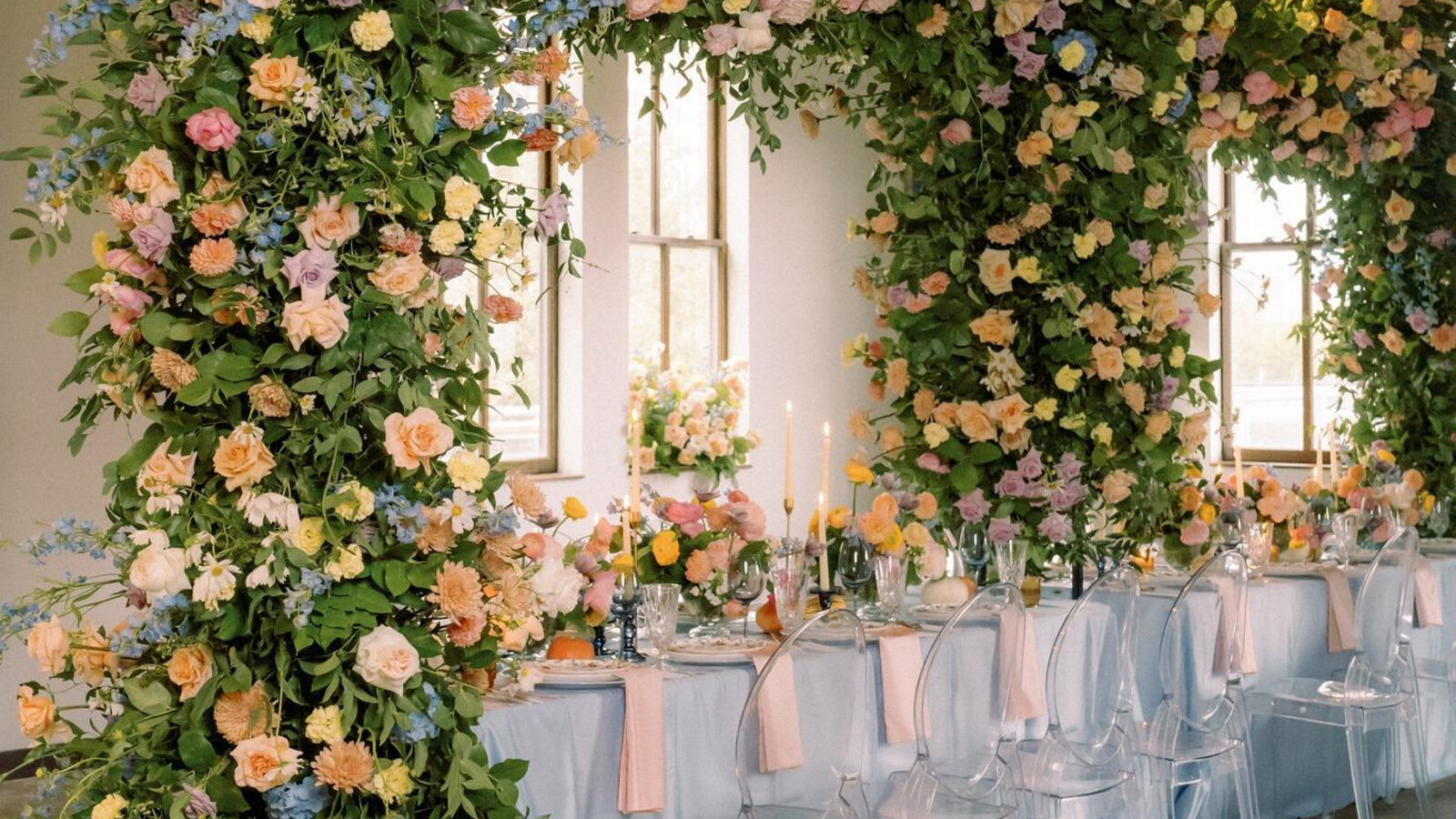 Elegant wedding reception with floral arch of peach, pink and blue flowers framing a table with clear chairs and star light above.