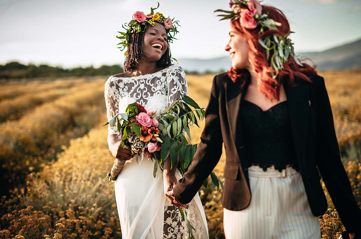 Two lesbian brides hold hands while walking in a flower field.