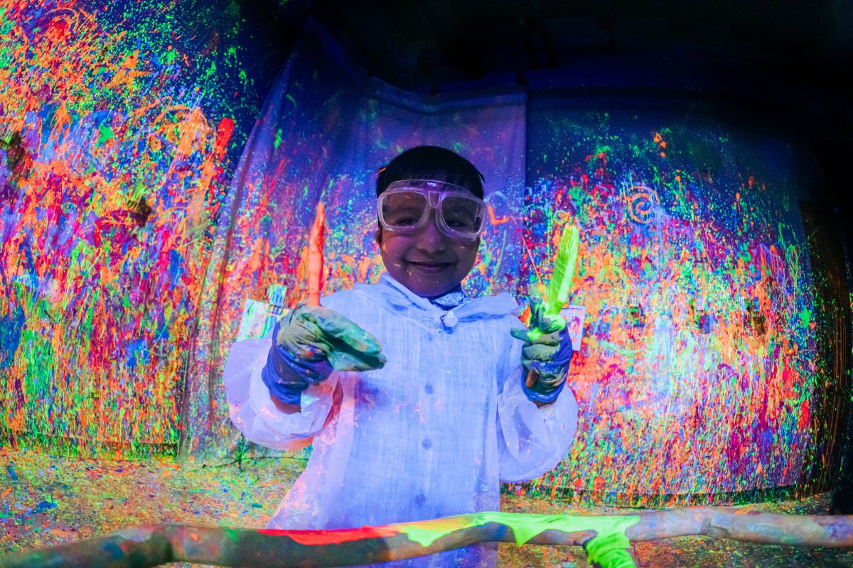 Smiling child holding paint brushes in the Blacklight Splatter Room at CAMP