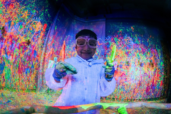 Smiling child holding paint brushes in the Blacklight Splatter Room at CAMP