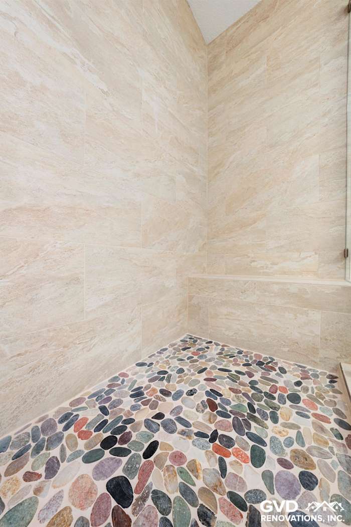 Custom tile shower with multi-colored pebble floor and cream stone wall tiles in Fair Oaks bathroom renovation