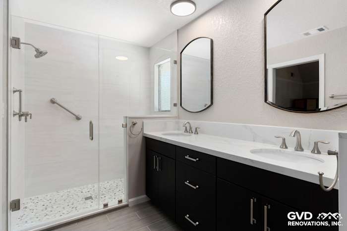 Modern master bathroom with dual sinks, dark cabinetry, glass shower, and light marble countertop