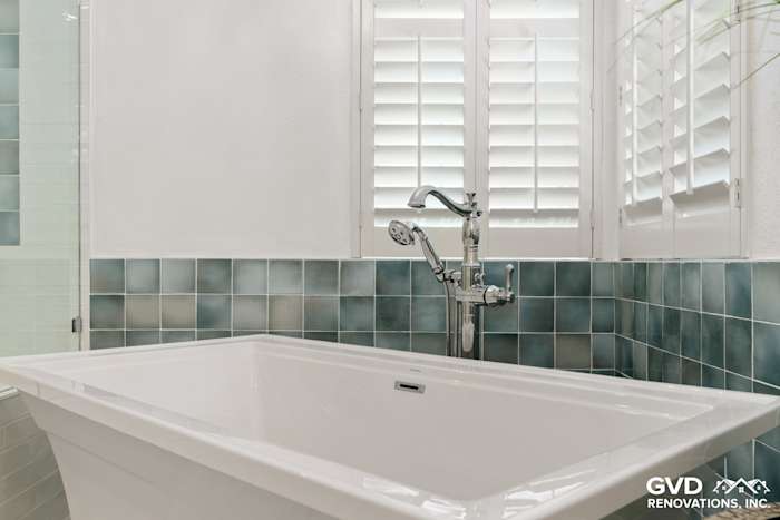 White freestanding bathtub with modern faucet surrounded by blue-green mosaic tile and window shutters in Fair Oaks bathroom