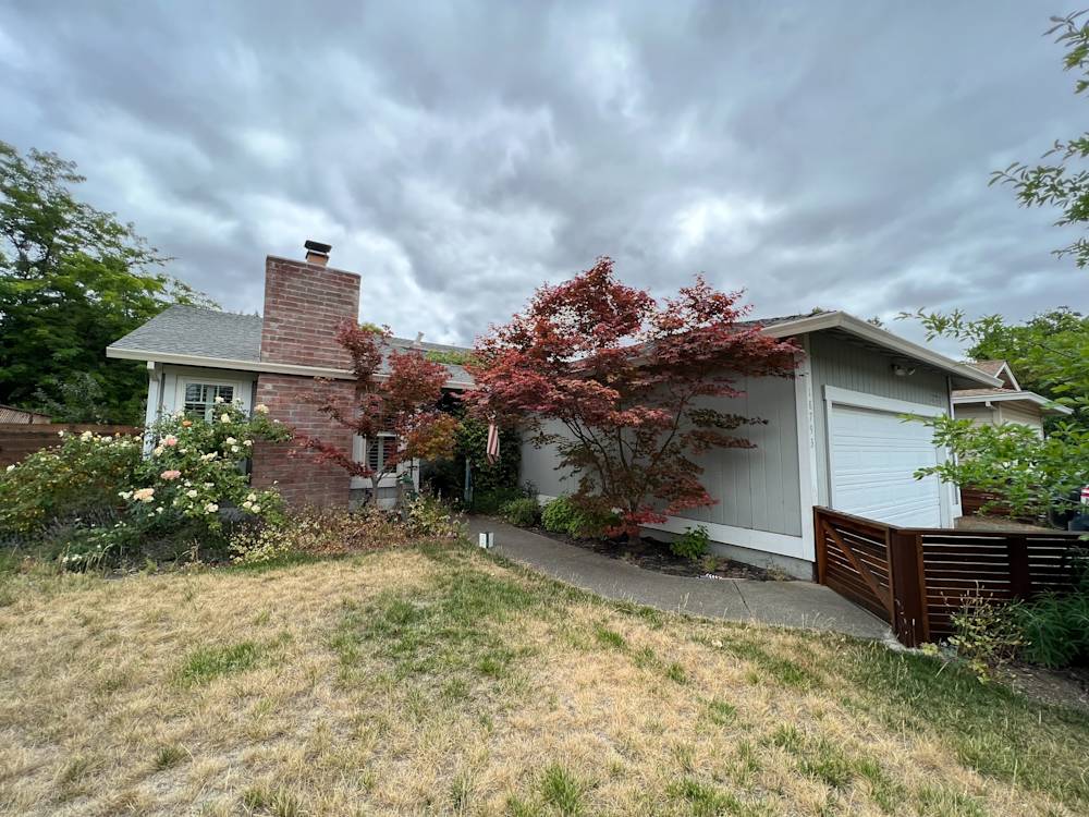 Charming Sacramento home with red brick chimney and overgrown shrubs, showing dry lawn and cloudy sky