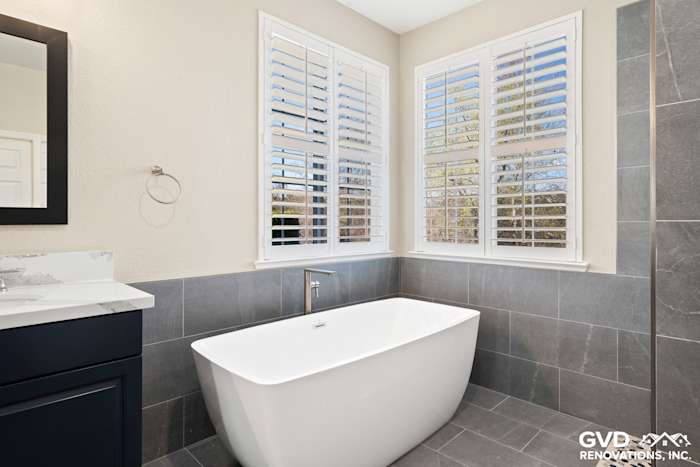 Modern master bathroom with freestanding soaking tub, gray tile, and natural light from white shutters