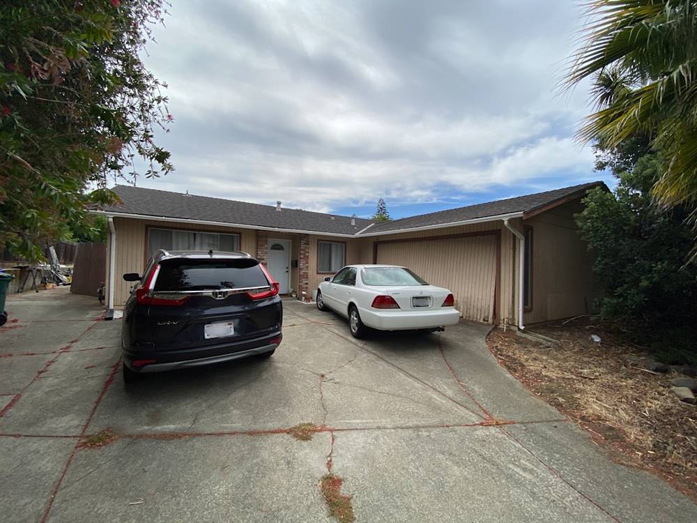 Single-story home in Mendocino with two cars parked on concrete driveway, overgrown landscaping, and neutral exterior