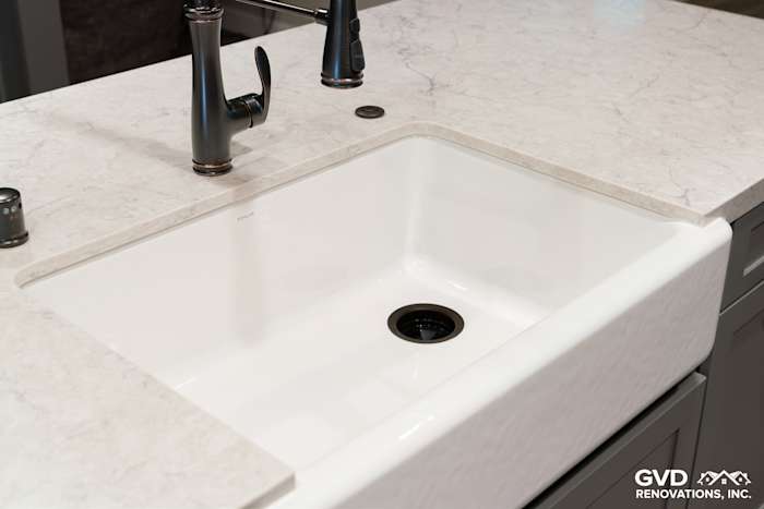 Modern kitchen sink with a white farmhouse basin and gray quartz countertop, featuring a sleek black faucet