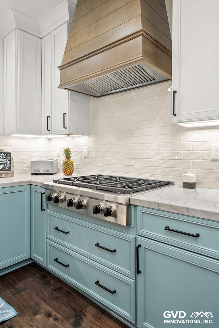 Bright kitchen with light blue base cabinets, white subway tile, and a wooden range hood above a gas stove