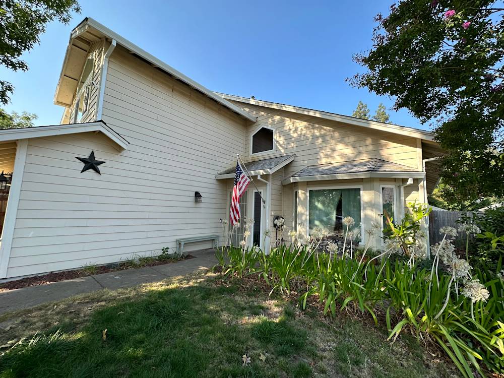 Dated exterior with beige siding, overgrown plants, and an American flag in Sacramento