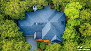 Aerial view of a beautifully roofed home featuring dark shingles, surrounded by lush greenery in a serene setting