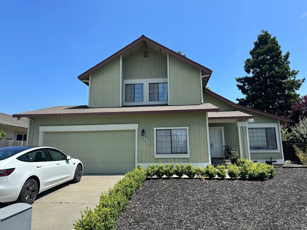 Two-story home with light green siding and gray garage, surrounded by dark mulch and manicured shrubs in Sacramento
