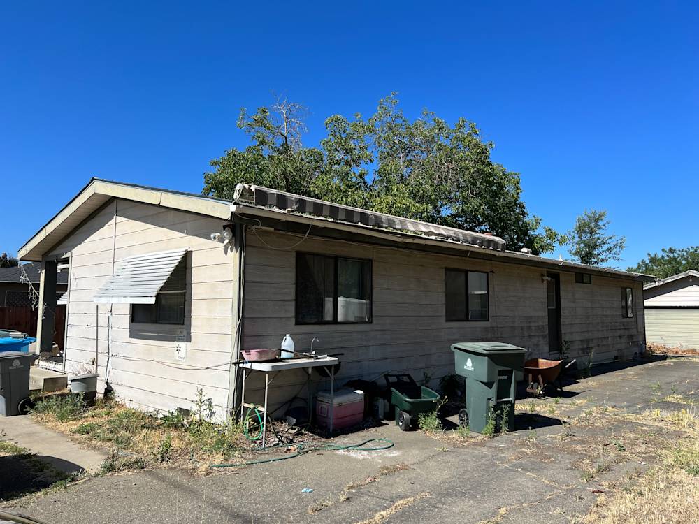 Single-story home with wood paneling, overgrown yard, and clear blue sky in Sacramento