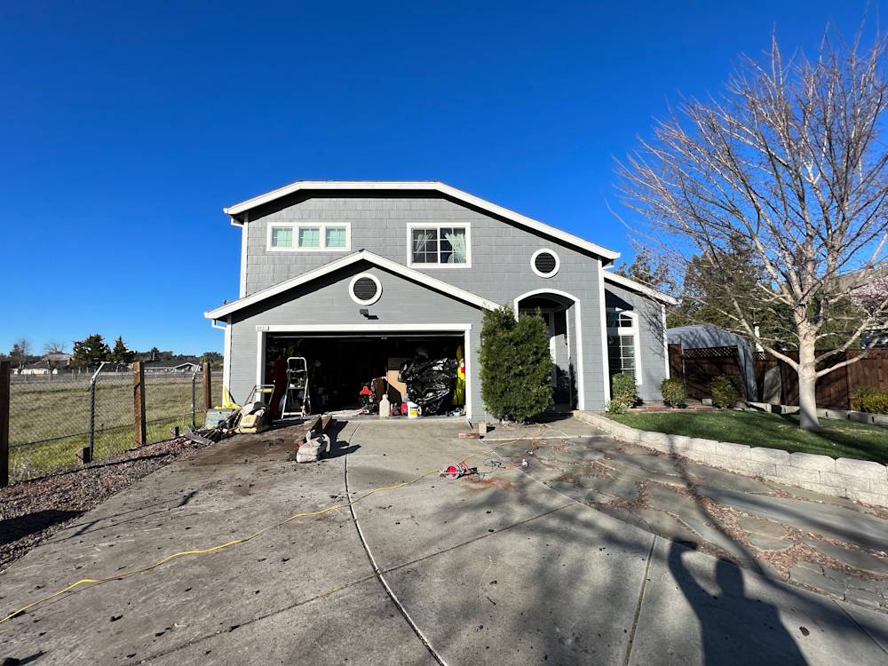 Gray two-story home with a concrete driveway and overgrown landscaping in Sacramento