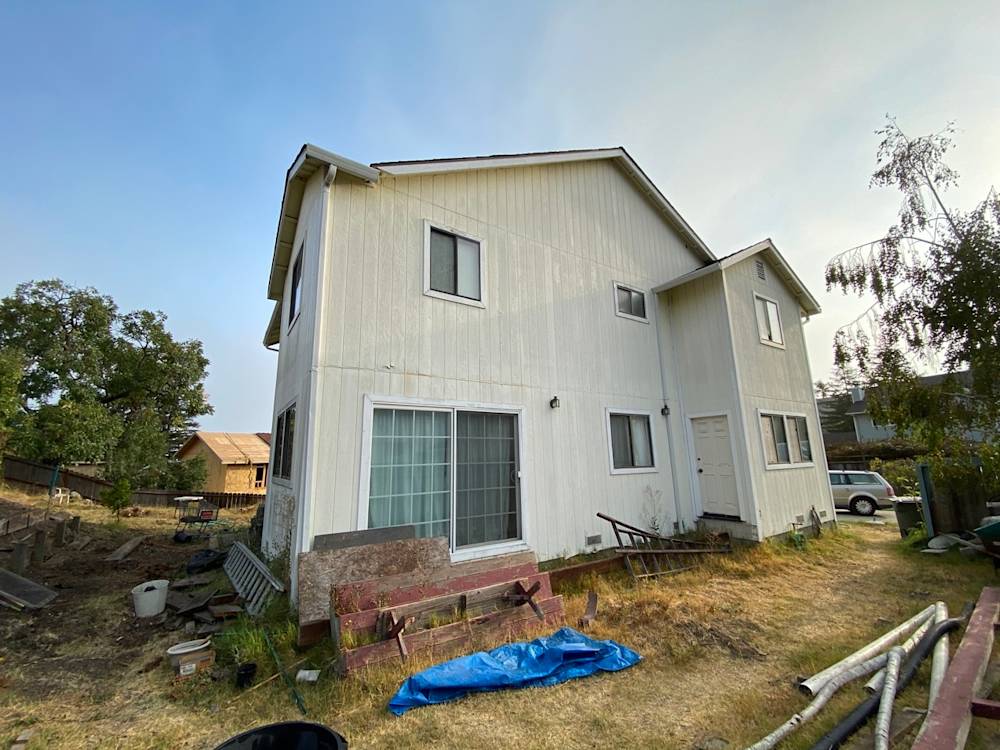 Side view of a weathered two-story home with white siding and unkempt landscaping in Northern California