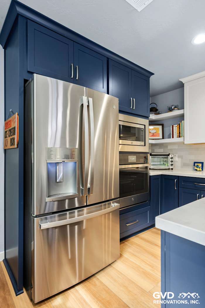 Modern kitchen with navy blue cabinetry, stainless steel refrigerator, double ovens, and light wood flooring