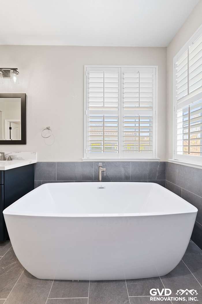 Modern master bathroom with freestanding soaking tub, gray tiled flooring, and natural light from white shutters
