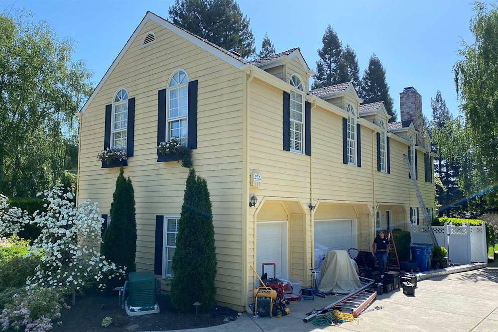 Wooden siding in pale yellow with dark shutters and outdated landscaping