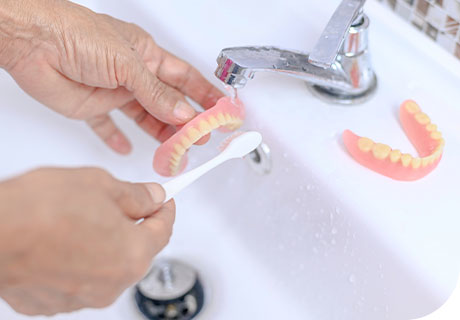 A close up of full dentures being cleaned with a tooth brush over a white sink. 