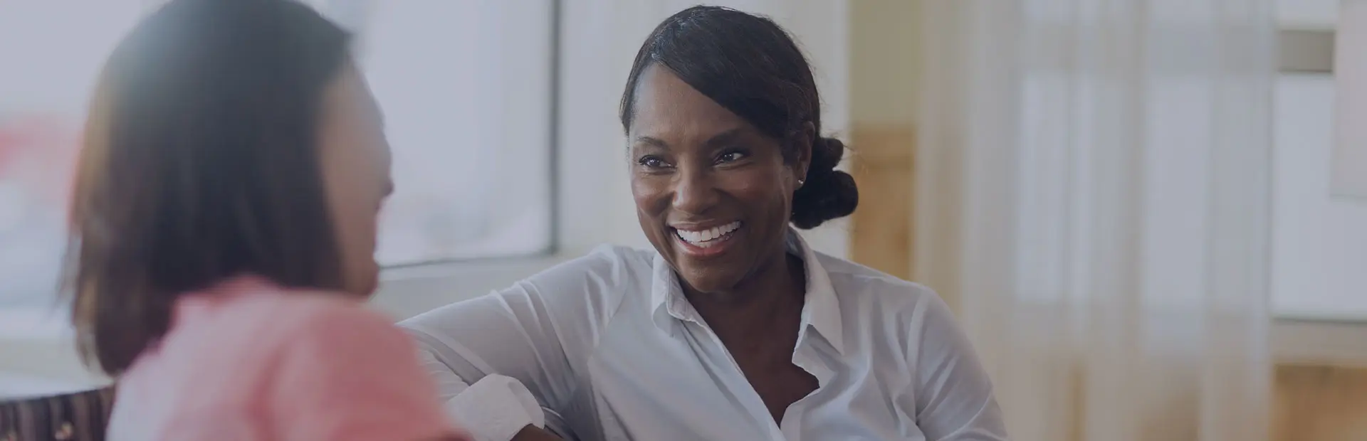 Two women are sitting on a striped sofa by the window having a conversation, one is smiling with a brilliant grin, confident she knows how to speak with dentures. 