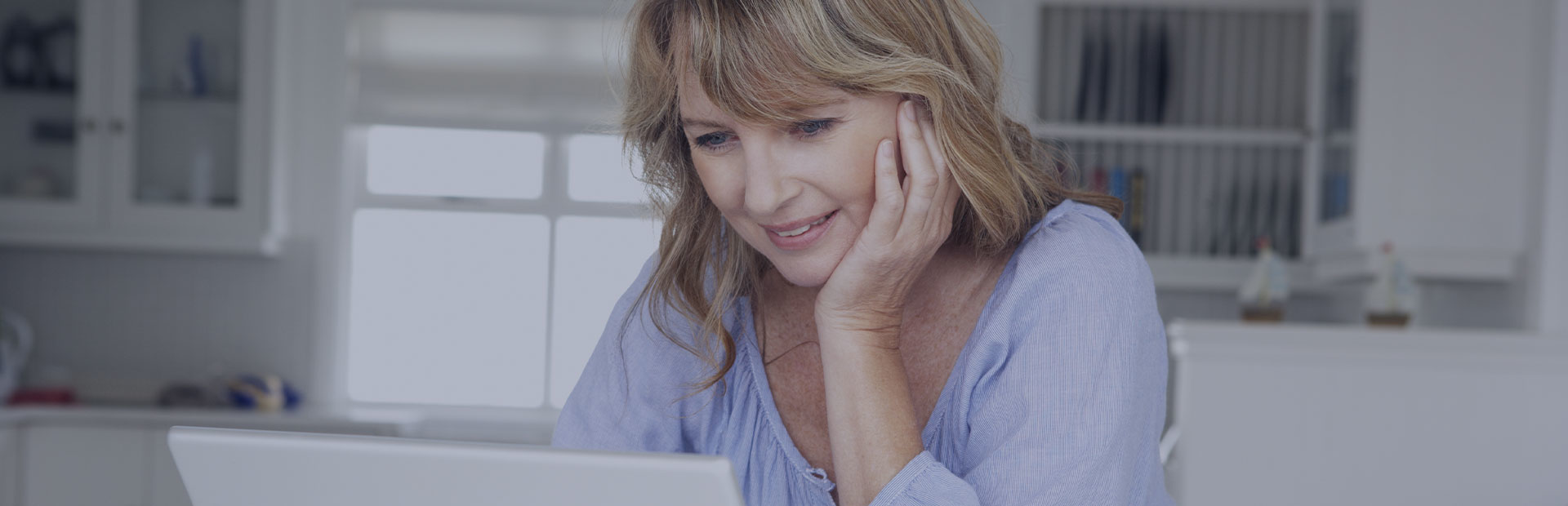 A woman in her early 50s is on her laptop in the kitchen, researching what she can do about denture pain and sores. 