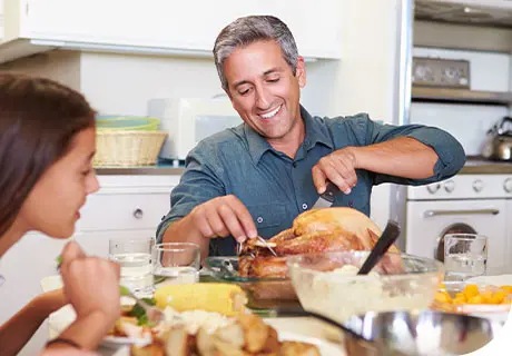 A man in his fifties is carving into a roast chicken, he is smiling because he knows he won't have any issues eating with dentures thanks to Fixodent's tips. 