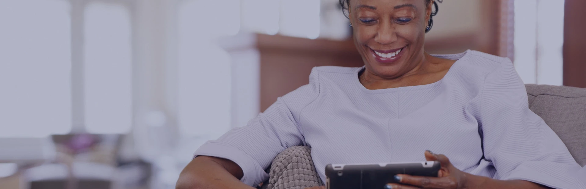 A woman in her 50s sits on a grey couch reading and iPad and is smiling, The text reads "Learn more about denture adhesive." 