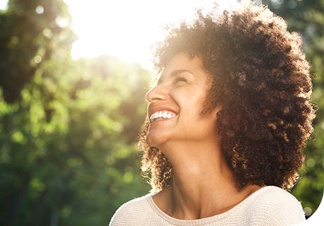 A woman in her forties is in a sunny garden and smiling as she thinks about choosing to get dentures or dental implants. 