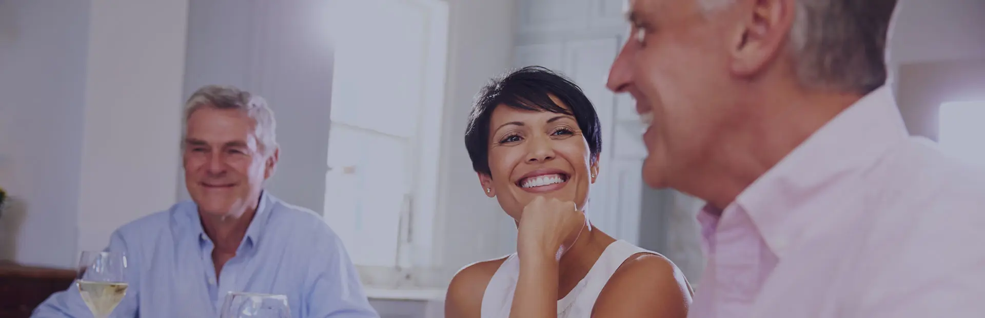 Two men and a woman in their fifties are sitting around the table chatting. The woman has a confident smile as she has no concerns about smiling with dentures.