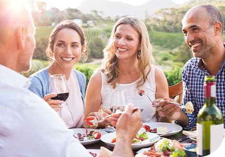 A group of friends in their forties are in a vineyard, dining al fresco, and they are all smiling, but you can't tell which one is wearing a type of partial denture. 