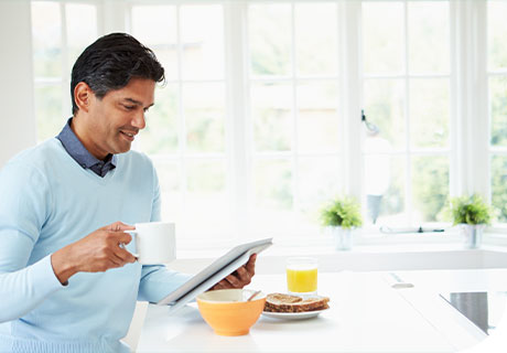A man in his 40s is sitting in a white kitchen drinking a coffee and having breakfast while reading a brochure on the costs of getting detures.