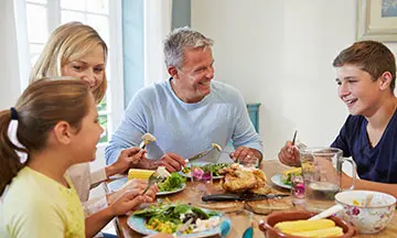A family sit around the table eating dinner, the father has dentures but is smiling confidently and eating meat and corn on the cob, as he knows about the benefits of denture adhesive cream.  