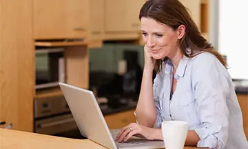 A woman in her 40s is in a wood-clad kitchen on her laptop reading the answers to the most frequently asked questions on Fixodent. 